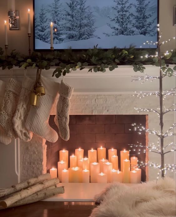a fireplace with pillar candles, branches, a lit up tree, white stockings and greenery on the mantel