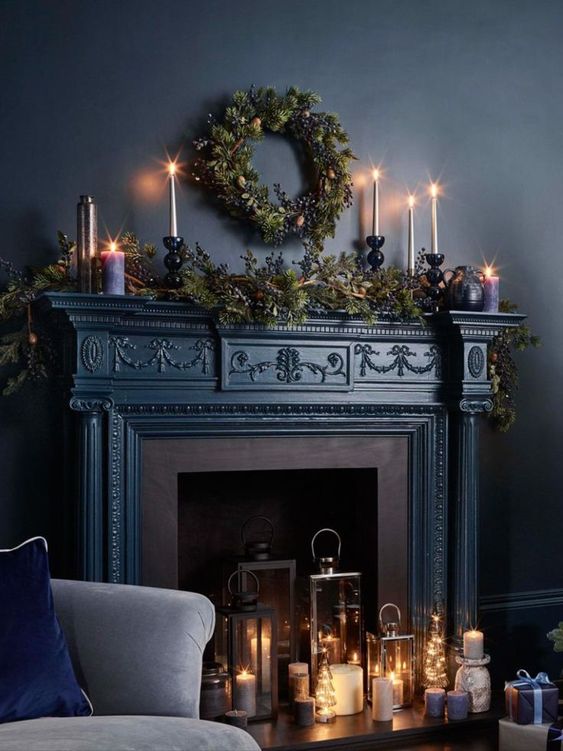 a gorgeous dark fireplace with candle lanterns and candleholders, evergreens and candles on the mantel