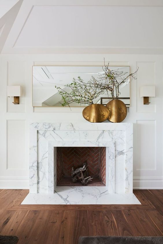 a large non-working fireplace clad with white marble and a mantel, with brass vases with green branches