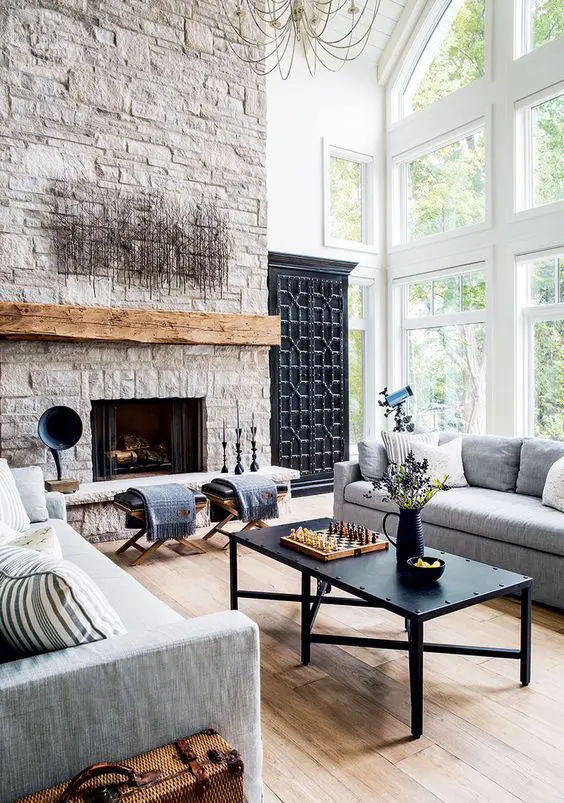 a living room with a stone fireplace, a stained mantel, stools, grey sofas, a black coffee table and some unique decor