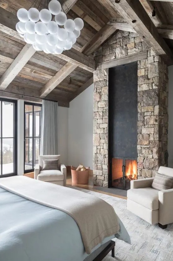 a modern chalet bedroom with a wooden ceiling, a bed with blue bedding, a stone clad fireplace and a bubble chandelier