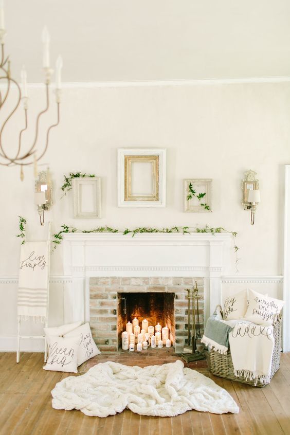 a neutral and cozy space with a fireplace with pillar candles, a rug, a basket with pillows, evergreens and empty frames