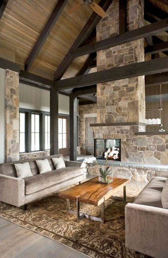 A neutral contemproary cabin living room with a stone double sided fireplace, neutral furniture, wooden beams
