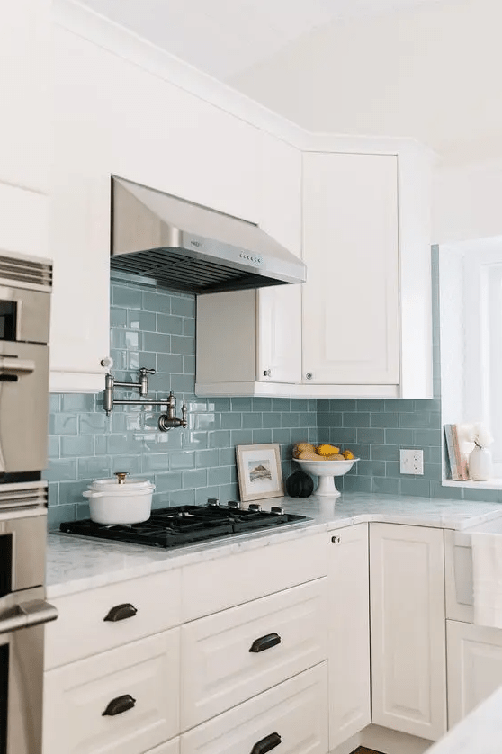 a neutral farmhouse kitchen with shaker cabinets, a blue subway tile backsplash and a metal hood, white stone countertops