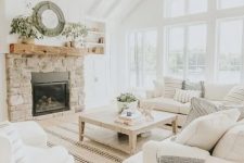 a neutral farmhouse living room with wooden beams, a round chandelier, white furniture, a built-in fireplace in stone