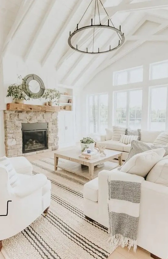A neutral farmhouse living room with wooden beams, a round chandelier, white furniture, a built in fireplace in stone