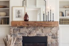 a neutral farmhouse space with a stone clad fireplace and a wooden mantel, candles, a mirror and some decor