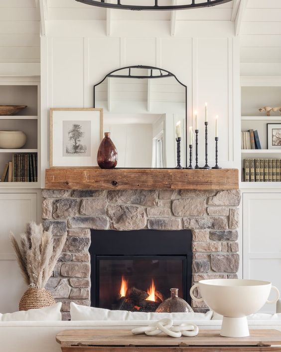 a neutral farmhouse space with a stone clad fireplace and a wooden mantel, candles, a mirror and some decor