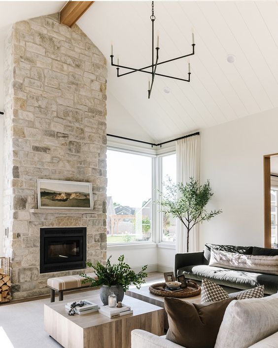 a neutral living room with a large stone clad fireplace, a dark green and creamy sofa, low coffee tables and greenery