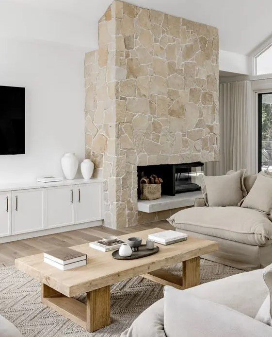 A neutral living room with a stone fireplace, neutral seating furniture, a low coffee table and white built in cabinets