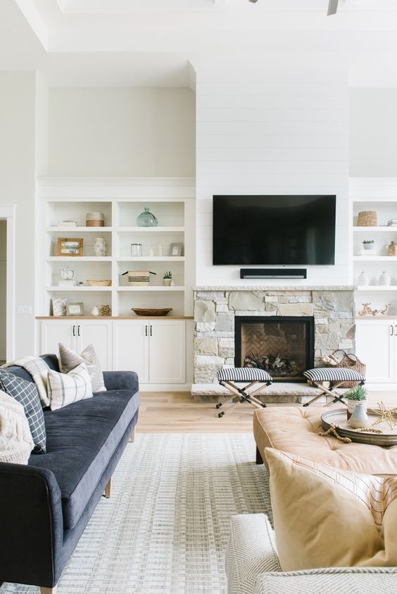 A neutral living room with built in storage units, a stone clad fireplace, a TV, a navy sofa, tan seating furniture