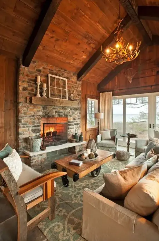 a welcoming chalet living room with a wooden ceiling and walls, a stone clad fireplace, modern and rustic furniture and an antler chandelier