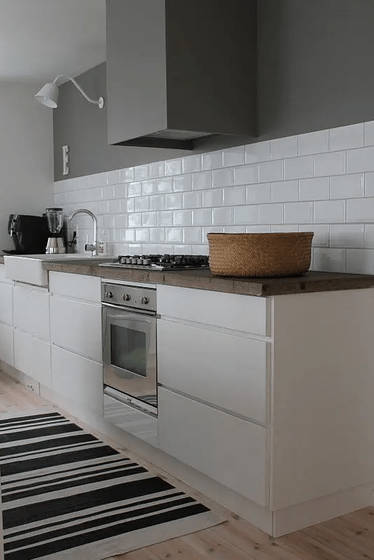 a white Scandinavian kitchen with sleek cabinets, butcherblock countertops, a white subway tile backsplash, a hood that matches the wall