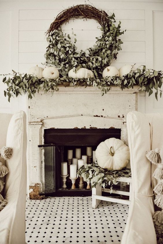 a refined neutral fall mantel with greenery, white pumpkins, a vine wreath with leaves, candles in the fireplace and a pumpkin on a chair
