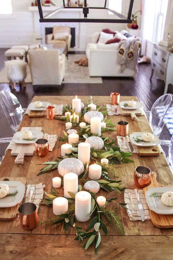 a modern rustic fall table setting with greenery, candles, neutral pumpkins, white plates on boards and copper mugs