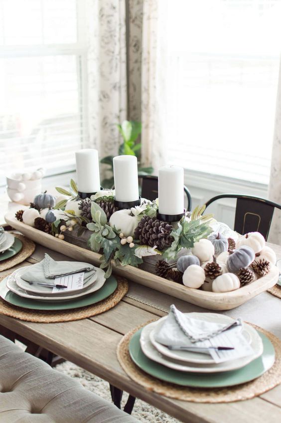 a stylish neutral fall tablescape with a wooden tray with candles, mini pumpkins, pinecones, greenery and berries, green and white plates and woven placemats
