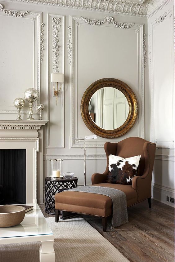 a very refined neutral living room with a brown wingback lounger, a refined round mirror and lots of molding on the walls