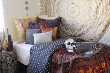 a boho Halloween bedroom with a mandala hanging, bold and dark bedding, bright sequin cushions, a metallic skull and black candles