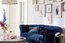 a bold and glam living room with a navy velvet Chesterfield sofa, a gallery wall, a printed round table a floral chandelier and bright blooms