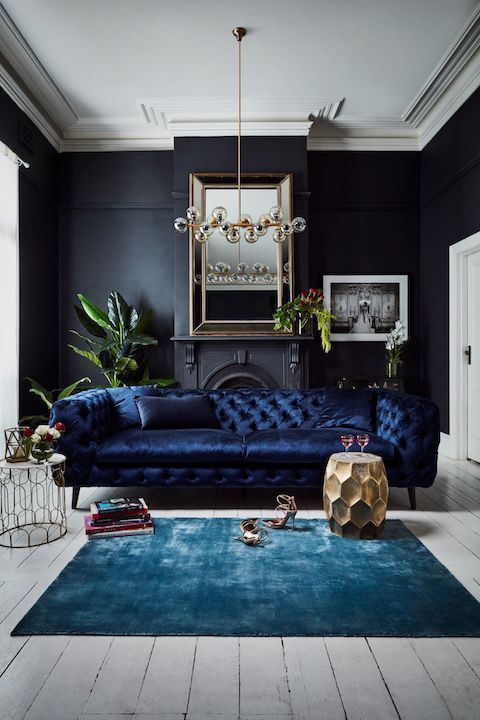 a classy and elegant living room with black walls, a navy velvet Chesterfield sofa, a blue rug, touches of gold