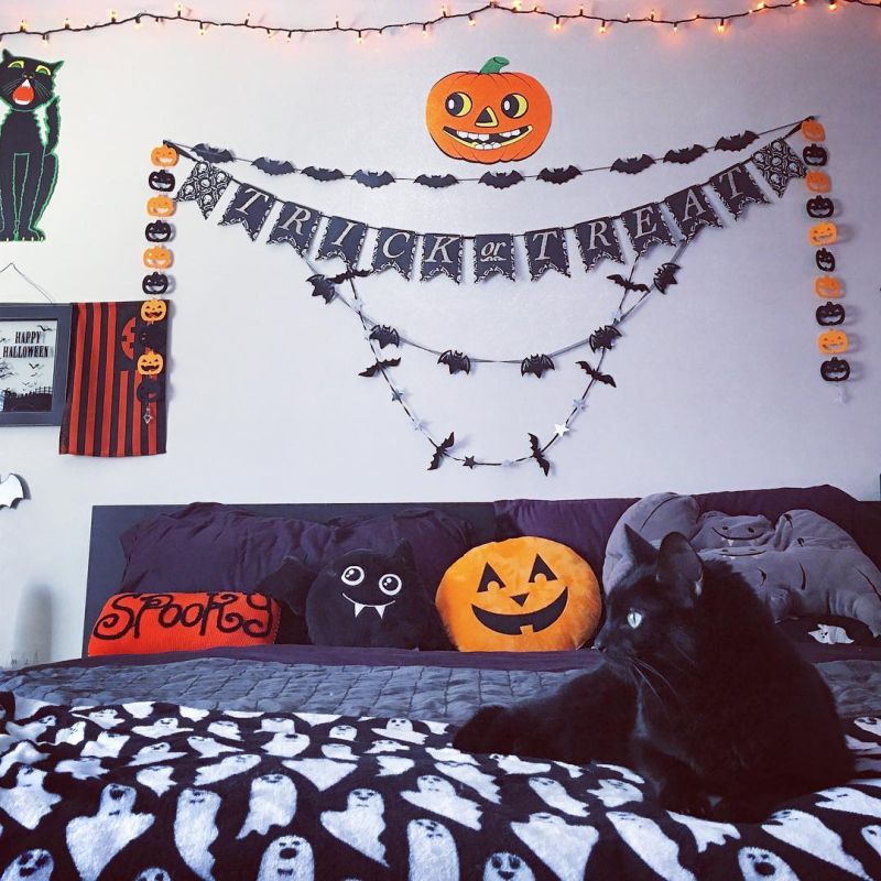 A fun and whimsy Halloween bedroom with bat and pumpkin garlands, lights, a black bed with fun pillows and plushies