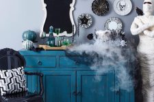a vintage blue console table with a white and turquoise pumpkin, an assortment of black and white decorative plates and smoke coming from the cauldron