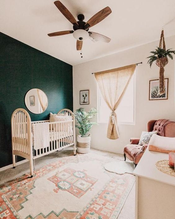 a welcoming gender neutral nursery with a boho rug, a dark green statement wall, a burlap curtain and greenery