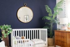21 a stylish boho nursery with a navy accent wall, a white crib and a wooden sideboard, a printed rug and a round mirror plus statement plants