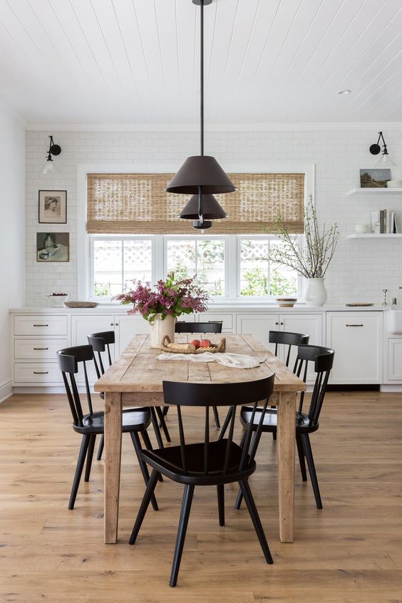 a cozy farmhouse kitchen with white lower cabinets, a rough wood dining table, black chairs and black pendant lamps