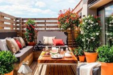a modern deck with a corner sofa, a wooden table, a candle lantern and lots of greenery and blooms in pots