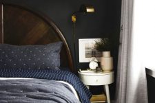 a moody bedorom with dark walls, a dark wooden bed, navy and blue bedding, a round nightstand and some greenery