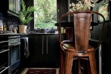 a moody kitchen in black, with black walls, cabinetry, a black tile backsplash, a boho rug, metal chairs and greenery and blooms