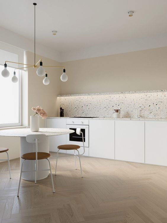 a neutral minimalist kitchen with a terrazzo backsplash, a round table, whimsy chairs and a minimal chandelier over the dining space