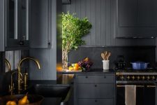 a stylish and refined moody kitchen with black cabinetry, a black backsplash, gold faucets for a chic touch