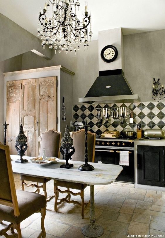 a vintage kitchen with black and white cabinets, a crystal chandelier, a wooden table, vintage chairs and candleholders plus a modern clock