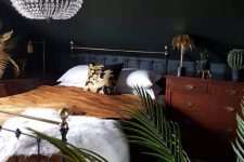 an elegant moody bedroom with a metal bed, a woodne nightstand, a statement crystal chandelier and potted plants