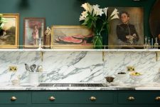 an elegant moody kitchen with hunter green walls and cabinetry, a marble backsplash, gold handles, a gallery wall on a ledge is very chic