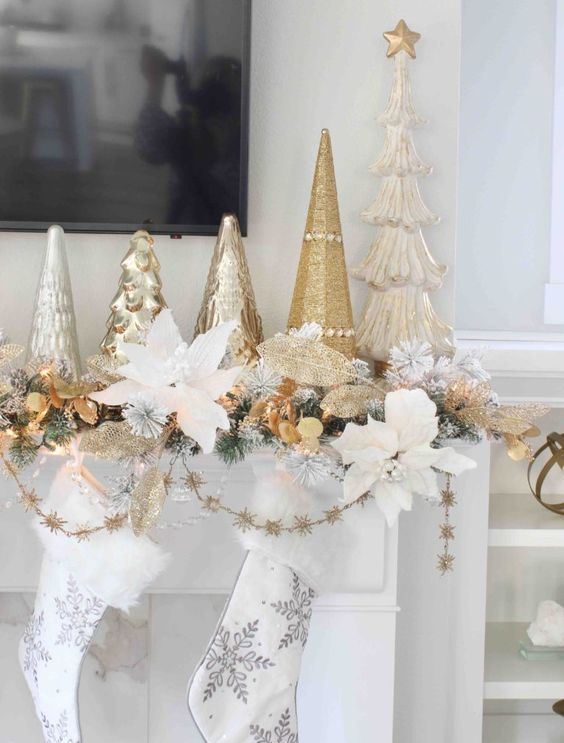 glam and chic Christmas mantel decor with snowflake stockings, gold and white flower garlands, leaves and an arrangement of Christmas trees