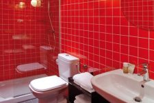 a bold modern red bathroom with medium size square tiles, white appliances, a round mirror, lights and white towels