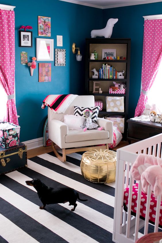 a bold nursery with navy walls, a striped rug, a gold ottoman, pink and white textiles and a bright gallery wall