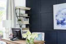 a chic rustic home office with a navy paneled wall, a wooden trestle desk, a basket, a white chair and a shelving unit plus a beaded chandelier