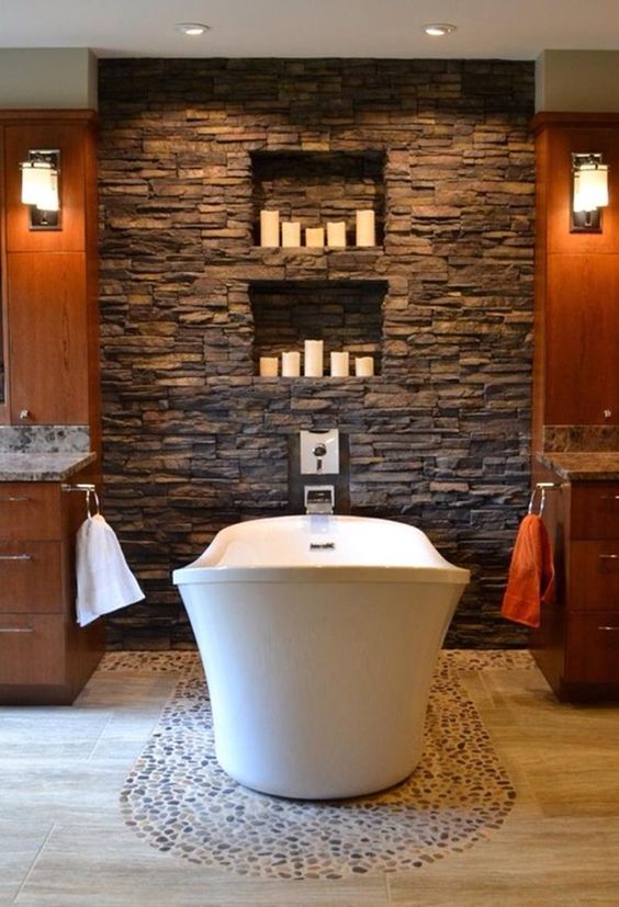 a cozy rustic bathroom with amber wooden cabinets, a faux stone wall with niches for candles and a mosaic pebble floor