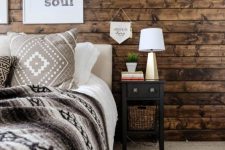 a farmhouse bedroom with a stained wooden wall, simple and cozy furniture, artworks and a table lamp and greenery