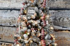 a flocked farmhouse Christmas tree with lights, plaid ribbons, a sign, a metal topper and some bells in various colors