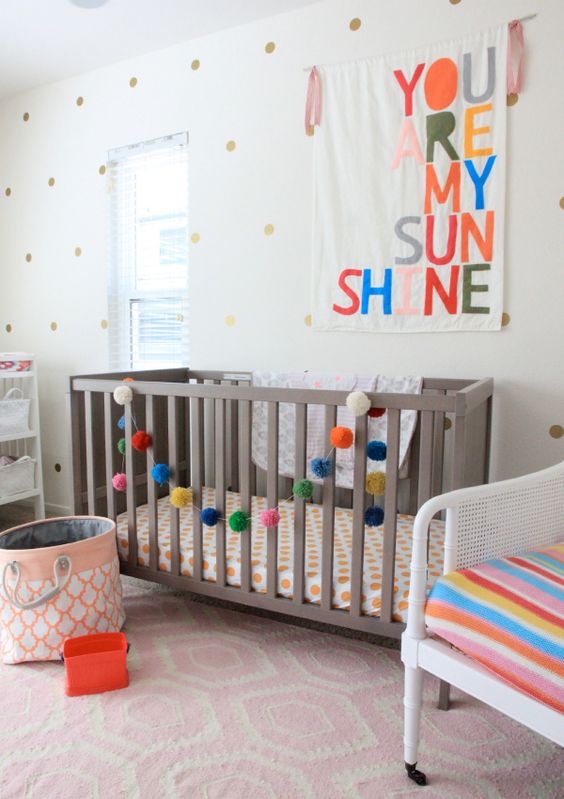 a fun nursery done with polka dot walls, a grey crib, white furniture, a pink rug, bright bedding, artworks and pompom garlands