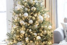 a gorgeoius and simple modern Christmas tree with white and silver ornaments of various sizes, lights and with a chunky knit cover