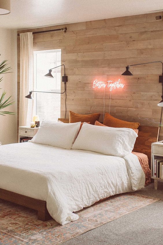 a modern bedroom with a whitewashed wooden wall, wooden furniture, sconces, statement plants and a pretty neon sign