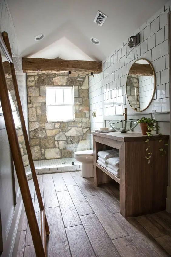 a modern rustic bathroom with a stone accent wall, wooden furniture, a round mirror and a glass bowl sink