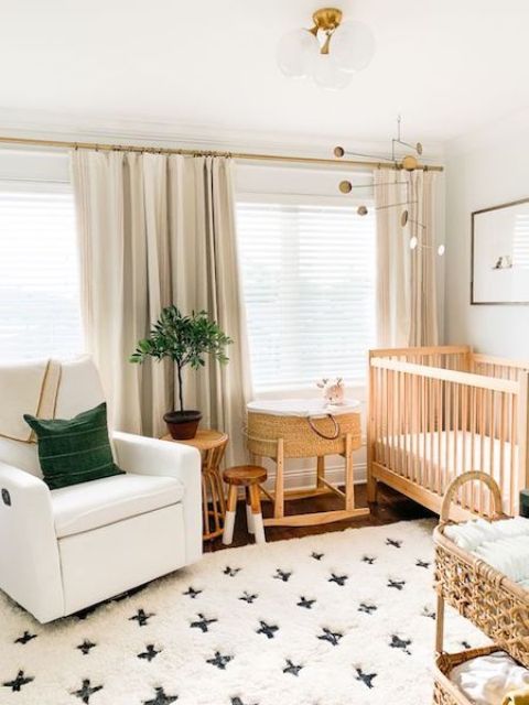 a neutral and cozy boho nursery with wooden furniture, a printed rug, a basket crib, neutral curtains and a potted plant