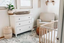 a neutral boho nursery with neutral and white furniture, printed textiles, a boho hanging on the wall, some potted plants and rugs